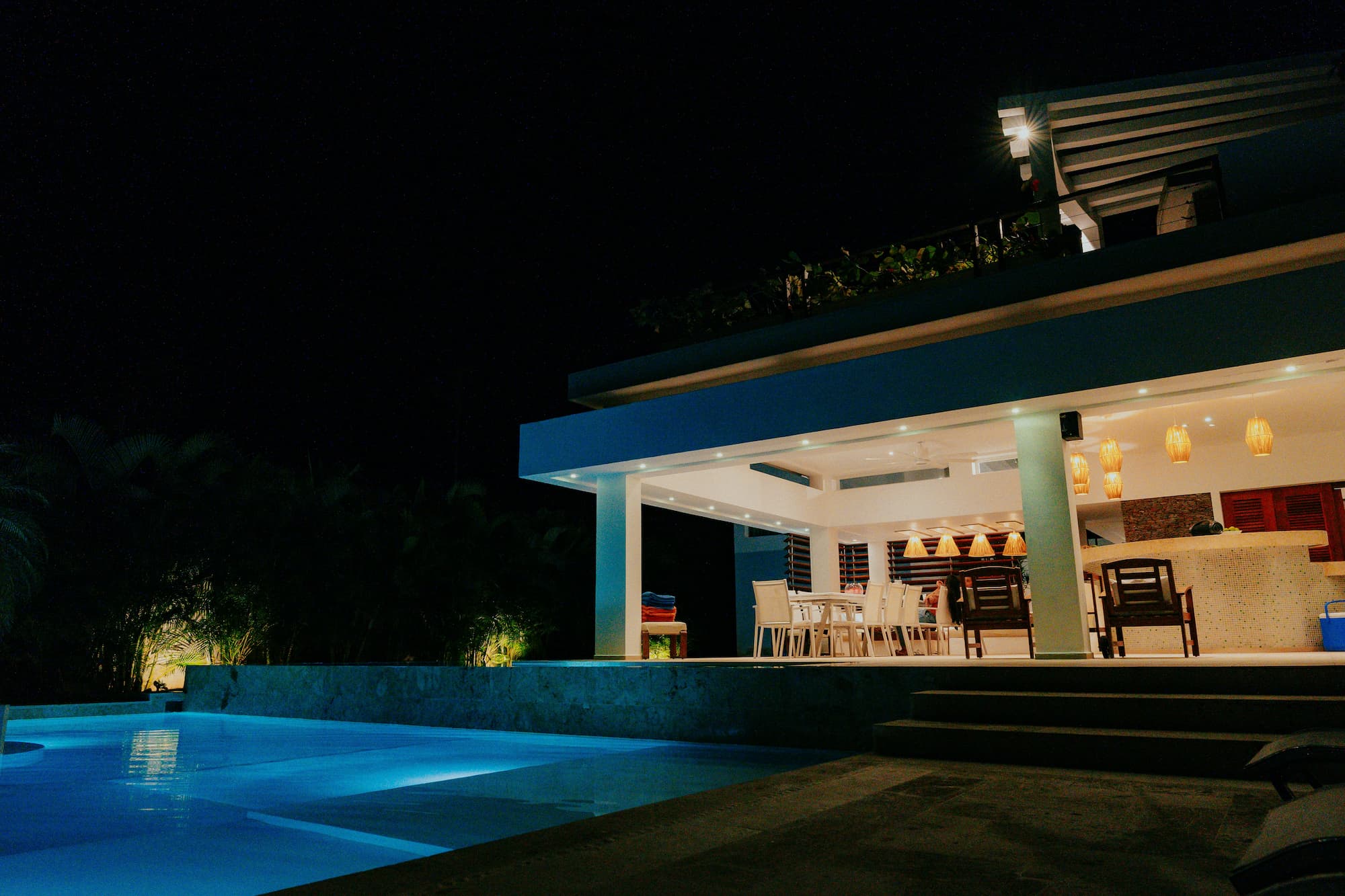 Een luxe villa met een verlicht zwembad en een overdekt terras met eettafel en bar, 's avonds gefotografeerd.